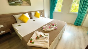 Een net opgemaakt bed met witte en gele kussens, een bloemstuk op het bed en een schilderij van een boslandschap aan de muur.
