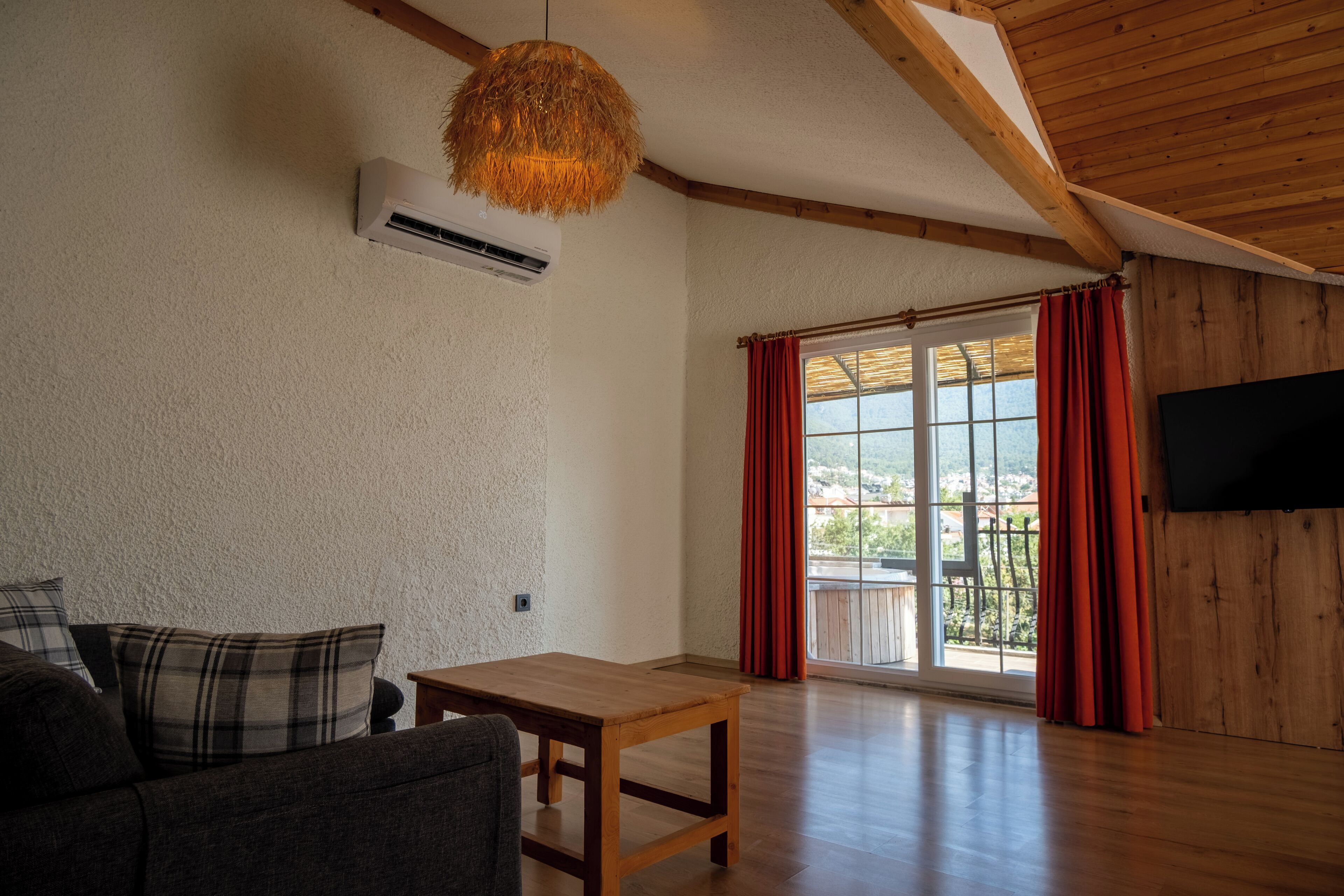 A spacious room with a wooden ceiling, a large window with red curtains, a flat-screen TV mounted on the wall, and a wooden table.