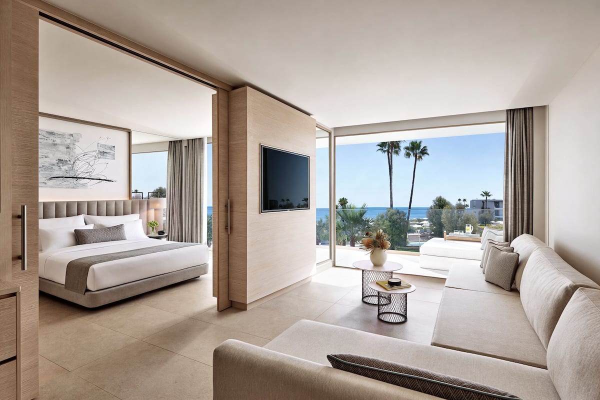 A modern hotel room with a bed, sofa, TV, and large windows with a view of palm trees and the sea.