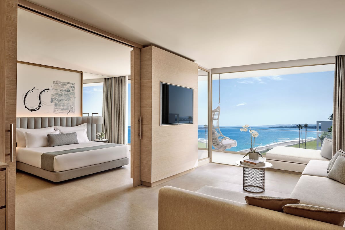 A modern hotel room with a bed, a sofa, a TV, and a large window with a view of the sea.