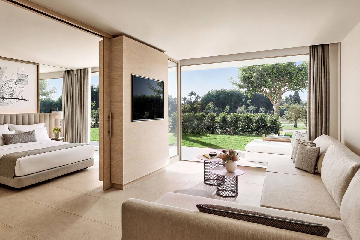 A modern hotel room with a large bed, a sofa, a TV, and a view of a garden.