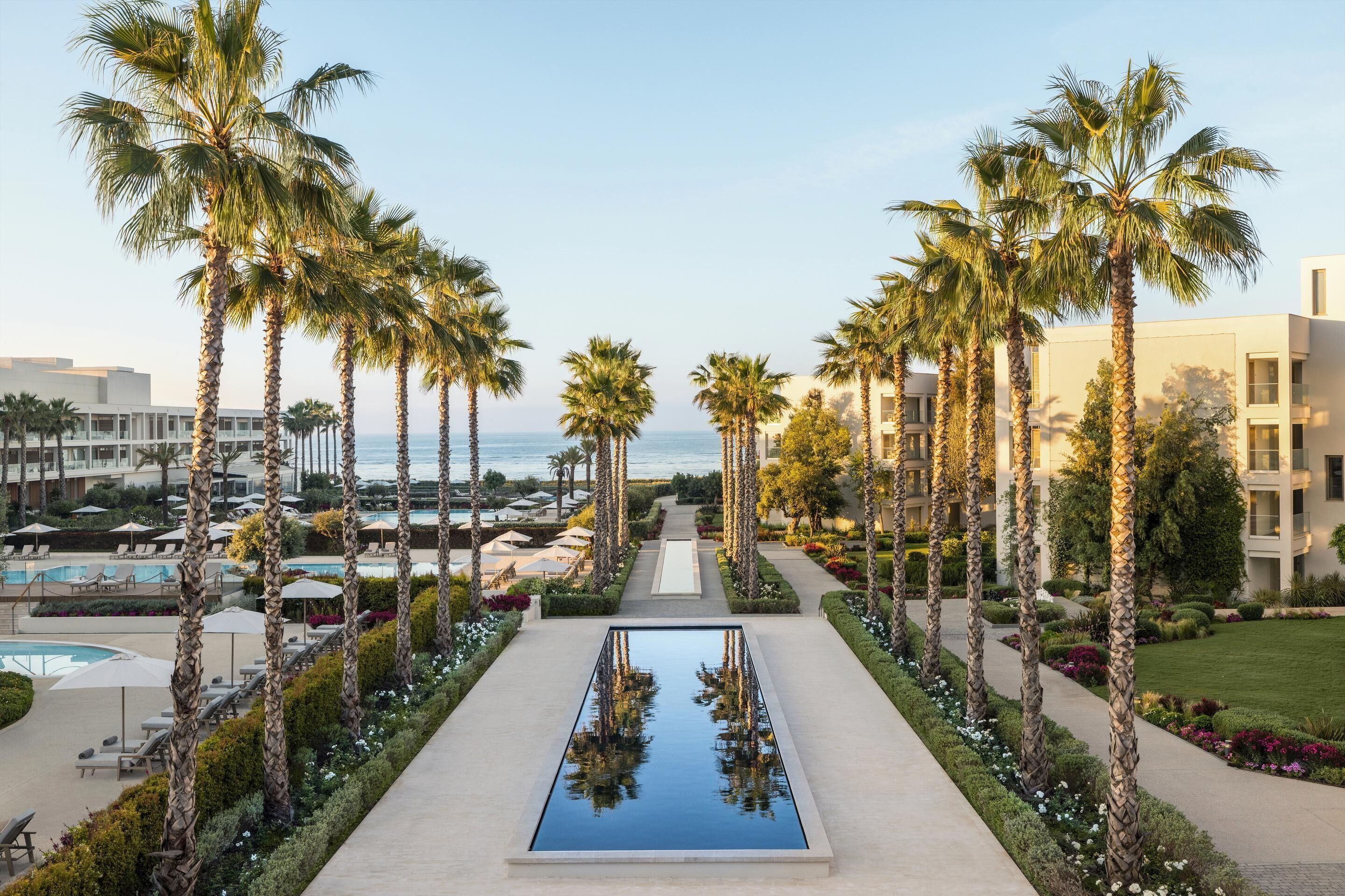 A resort with a central pool flanked by palm trees, a poolside area, and a view of the ocean.