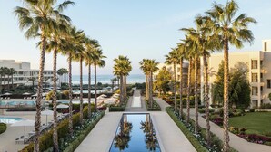 A resort with a central pool flanked by palm trees, a poolside area, and a view of the ocean.