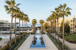 A resort with a central pool flanked by palm trees, a poolside area, and a view of the ocean.