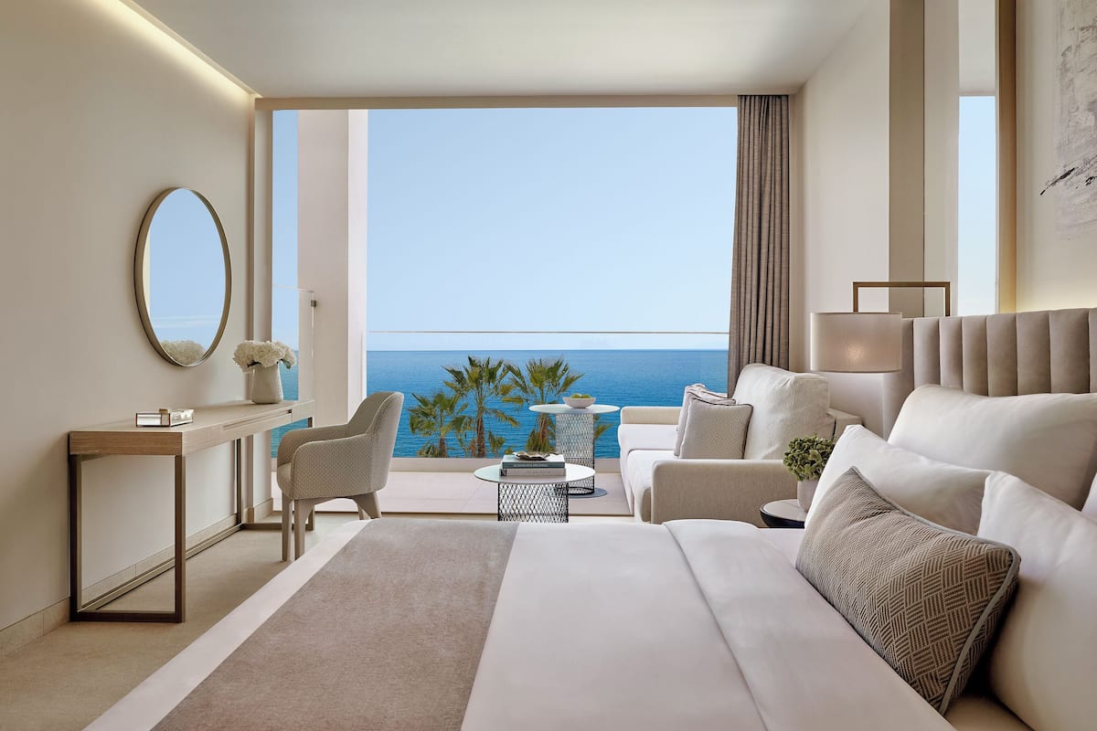 A modern hotel room with a large bed, a desk, a chair, a small round table, and a view of the ocean.