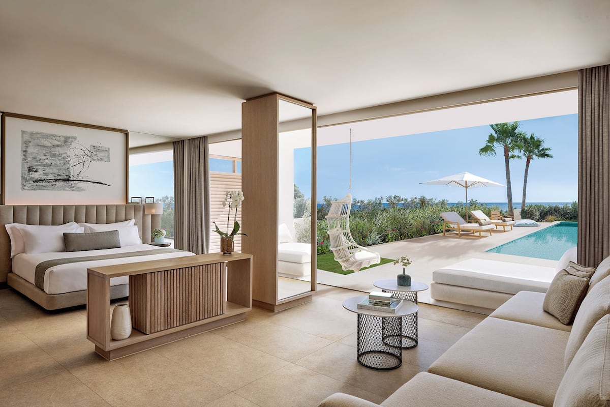 A modern hotel room with a large bed, a sofa, a coffee table, and a view of a pool and outdoor seating.