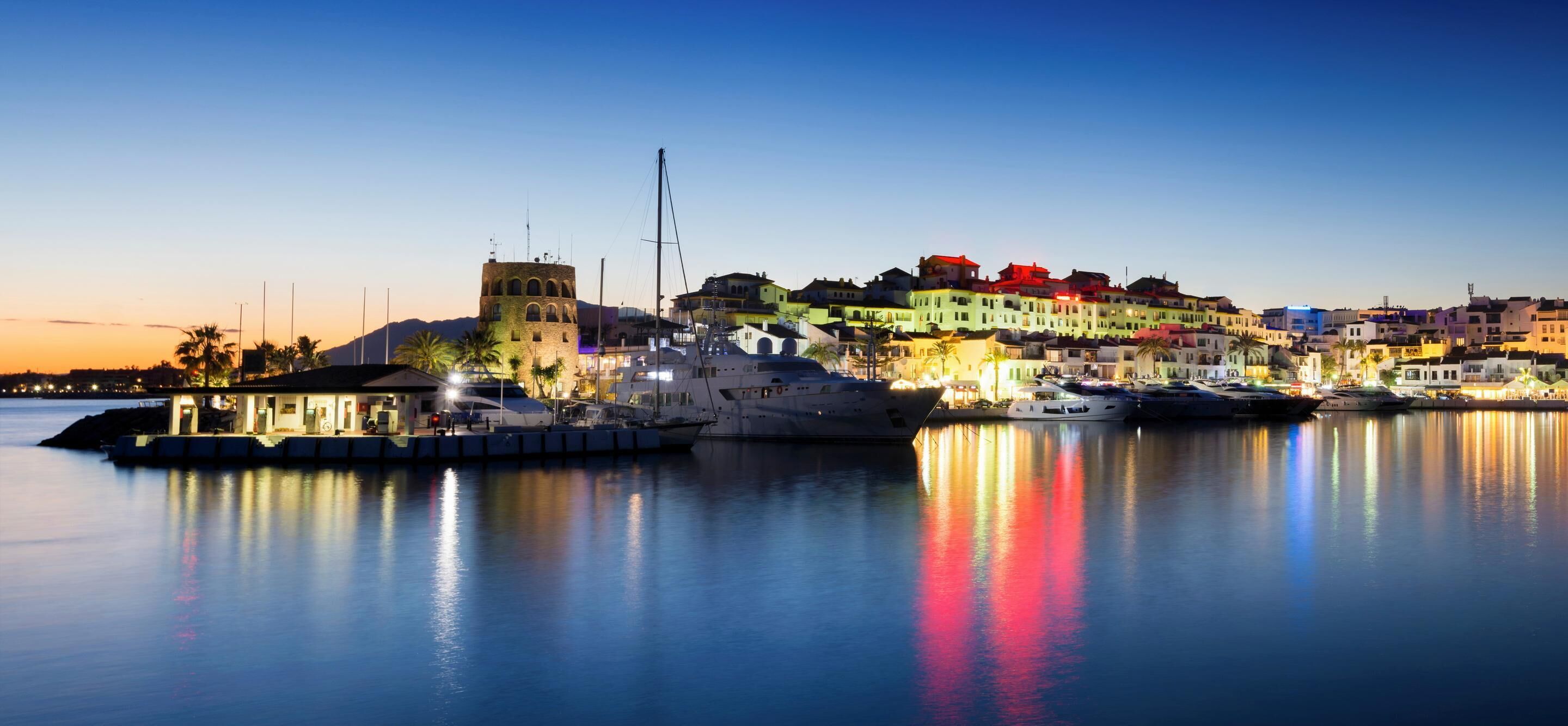 A waterfront marina with boats, a prominent tower, and colorful buildings along the shore.