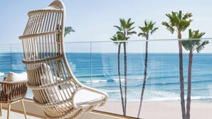 A balcony with a wicker chair, a small table, and a view of the ocean and palm trees.