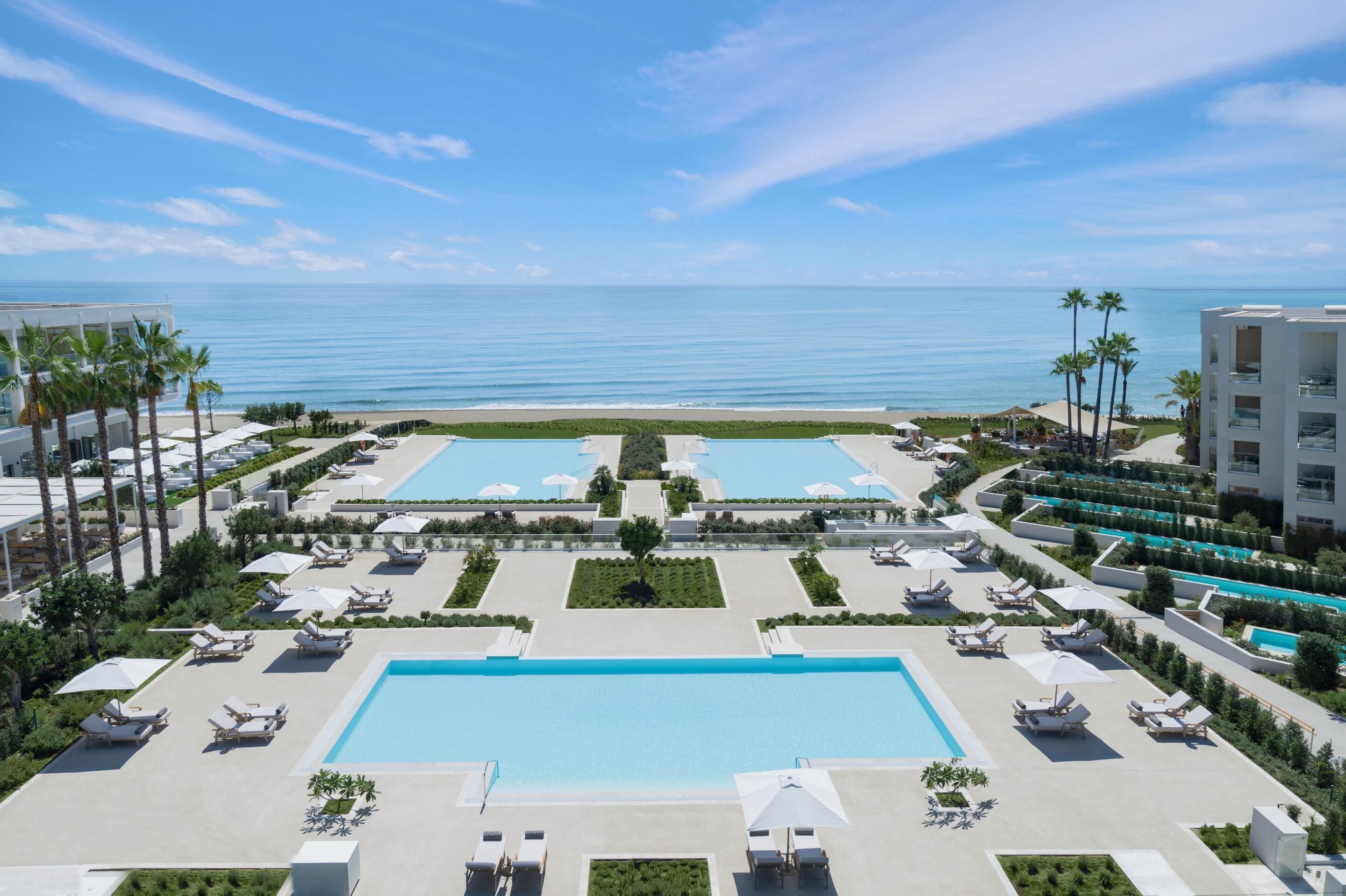A resort with two large swimming pools, sun loungers, palm trees, and a view of the ocean.