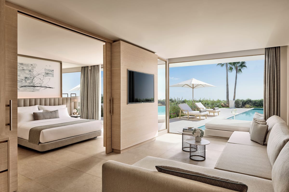 A modern hotel room with a bed, a sofa, a TV, and a view of a pool and palm trees.