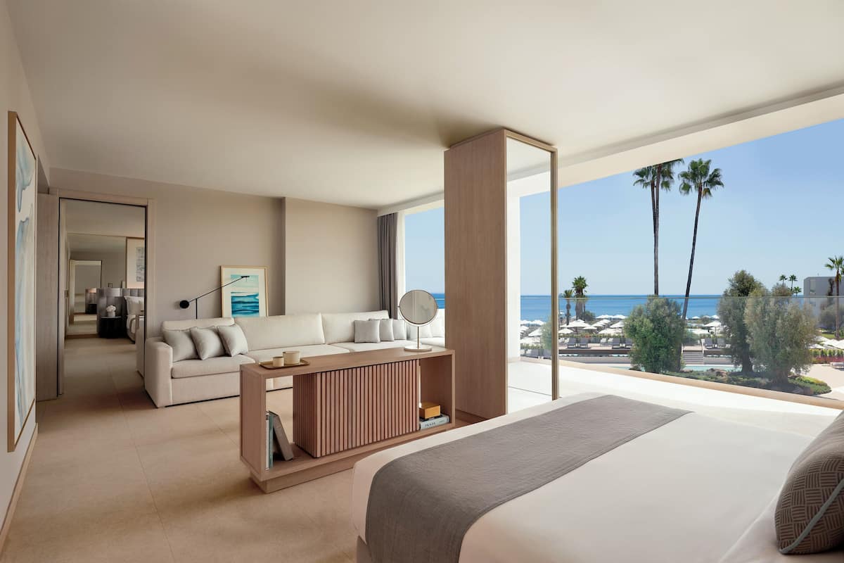 A modern hotel room with a bed, a sofa, a wooden console table, a large window with a view of the beach and palm trees.