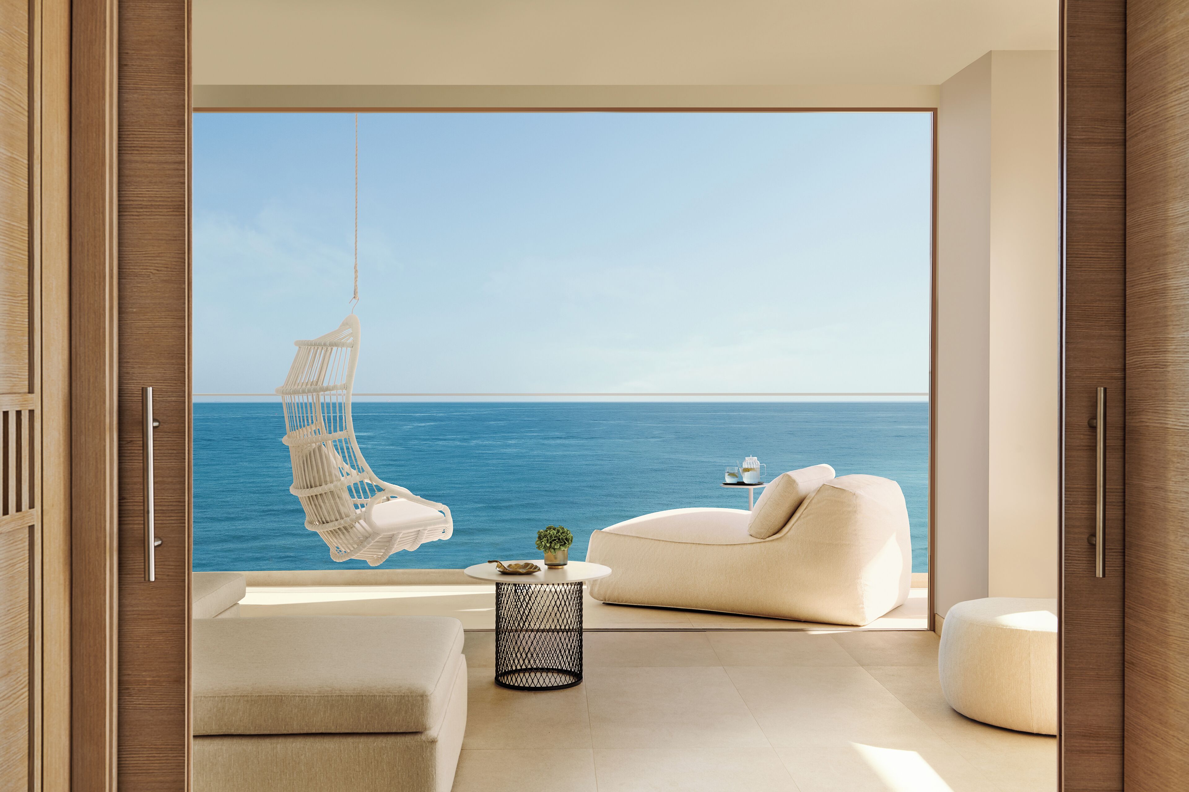 A modern interior with a large window overlooking the sea, a hanging chair, a round table with a plant, and a sofa.