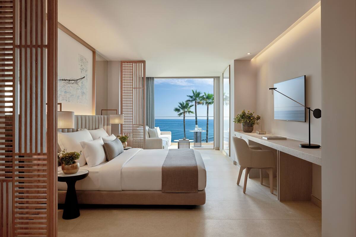 A modern hotel room with a bed, desk, and large windows overlooking the sea.