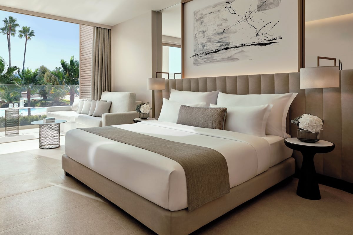 A modern hotel room with a large bed, two bedside tables, a sofa, and a balcony with a pool view.