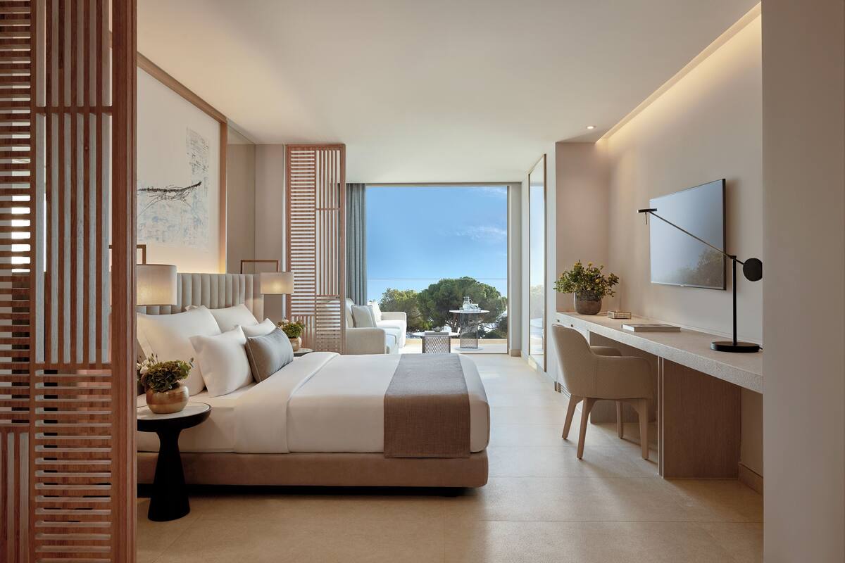 A modern hotel room with a bed, desk, and large windows.