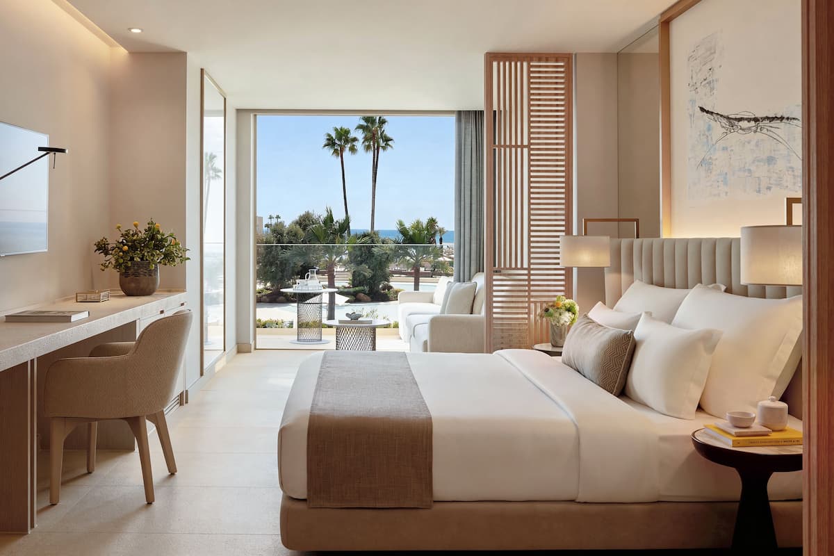 A modern hotel room with a large bed, a desk, a chair, a balcony with a view of palm trees and the ocean.