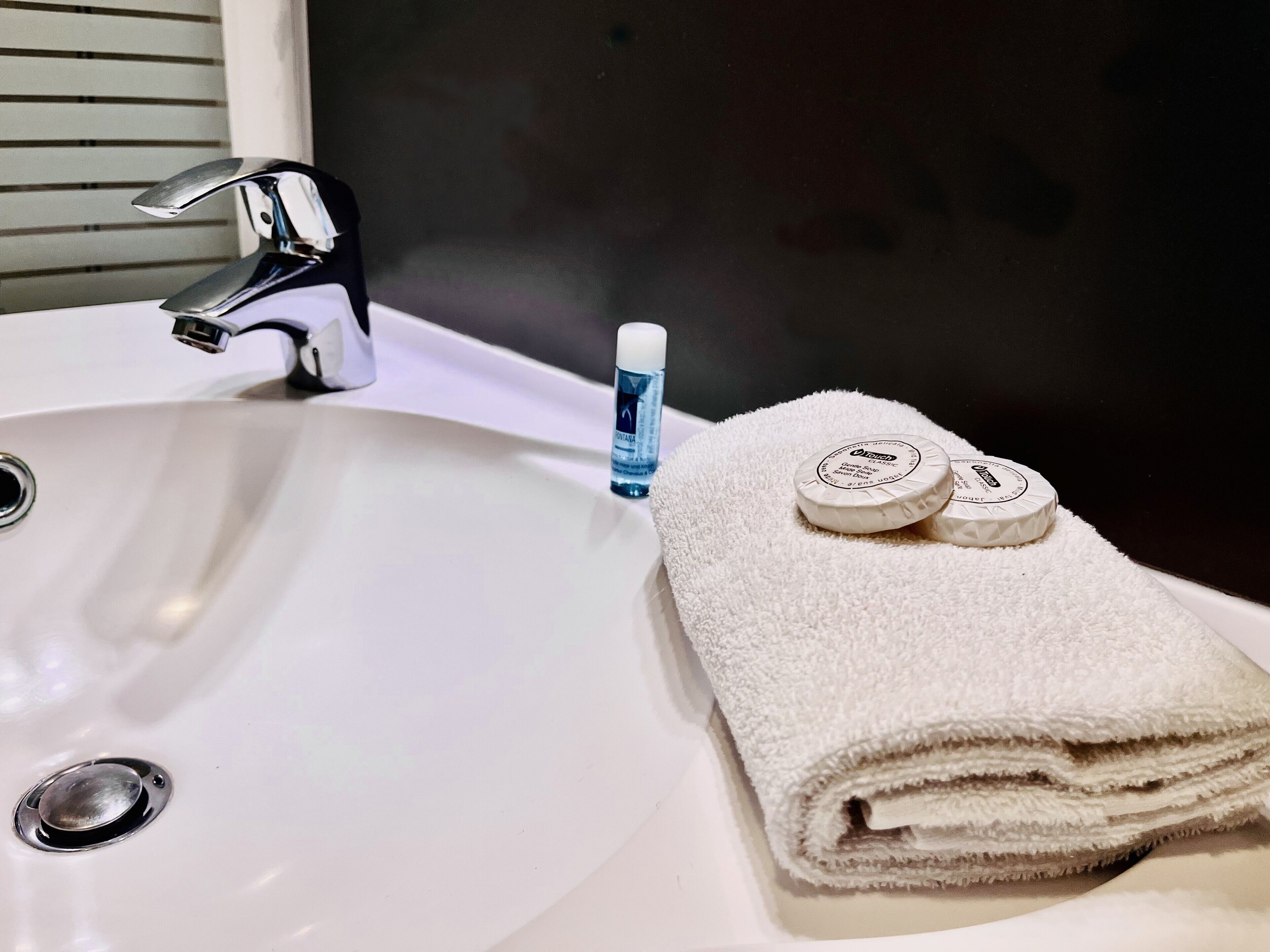 Un lavabo blanc dans une salle de bain, doté d’un robinet chromé, une bouteille bleue de savon pour les mains et une serviette blanche pliée ornée d’un logo circulaire.