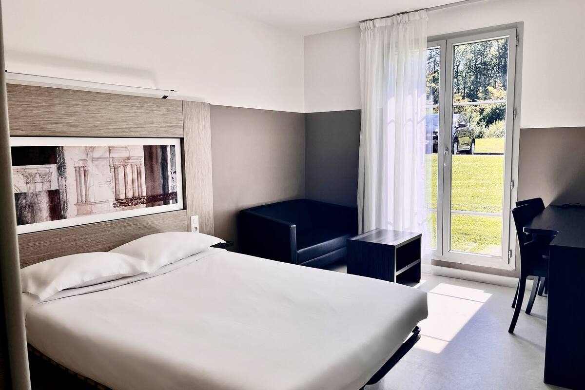 A hotel room with a bed, a desk, and a chair. There is a window with a view of a green landscape.