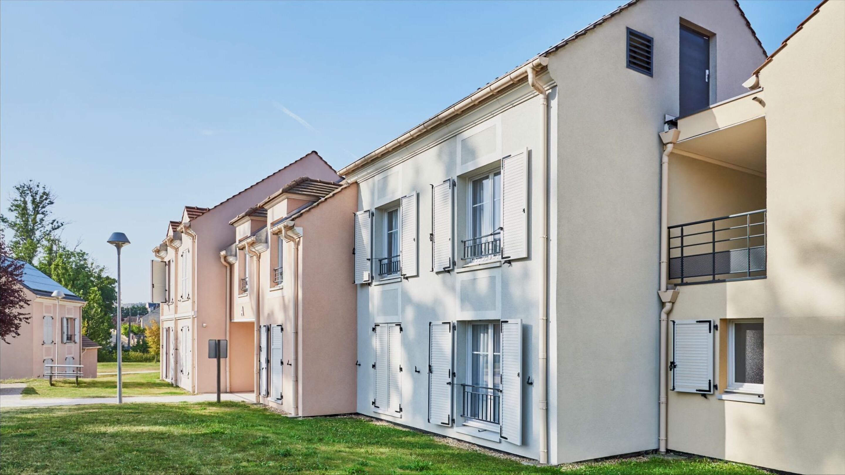 A row of modern apartment buildings with balconies, a well-maintained lawn, and a clear sky.