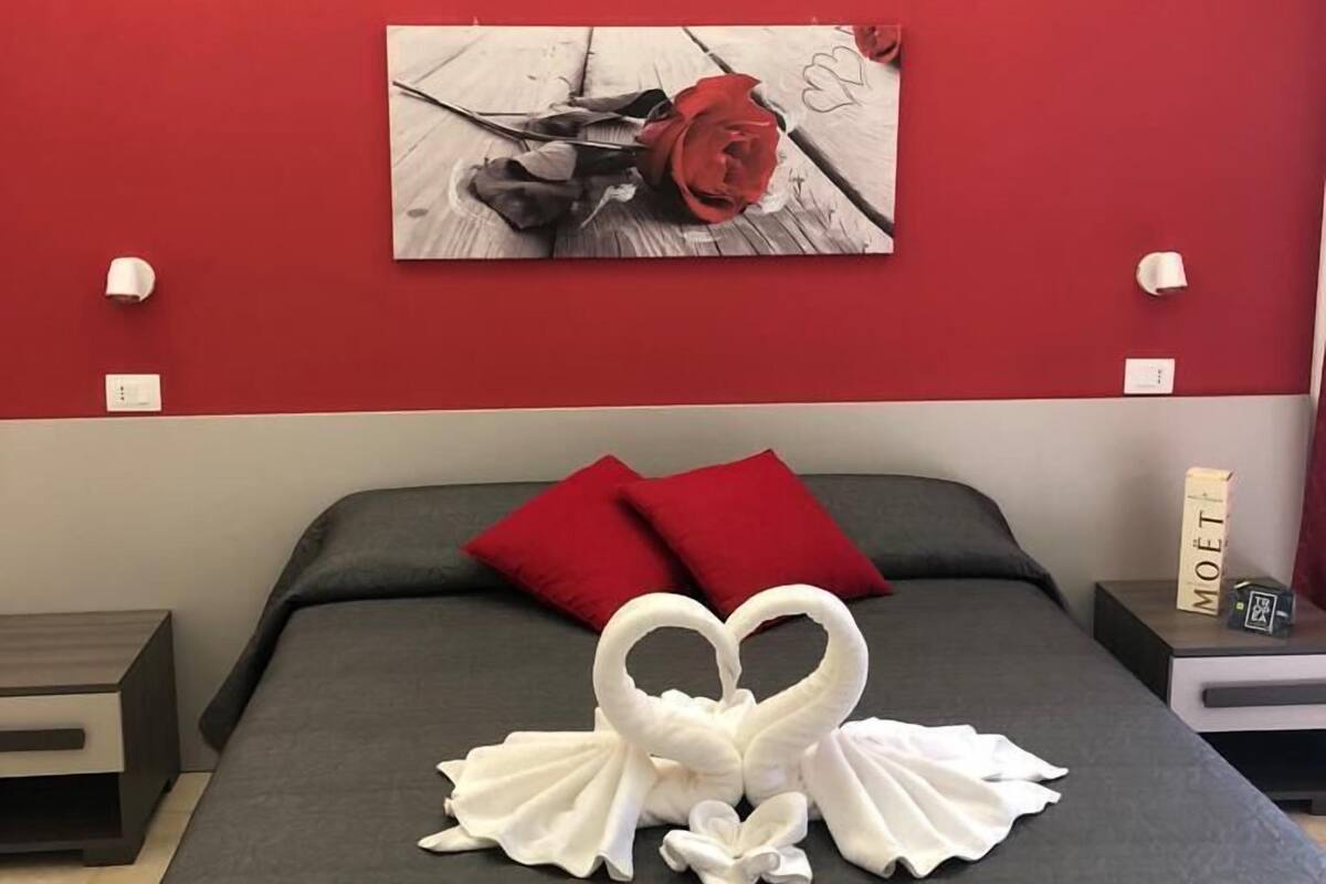 A hotel room with a red accent wall, a bed with a heart-shaped towel arrangement, and a picture of a red rose on the wall.