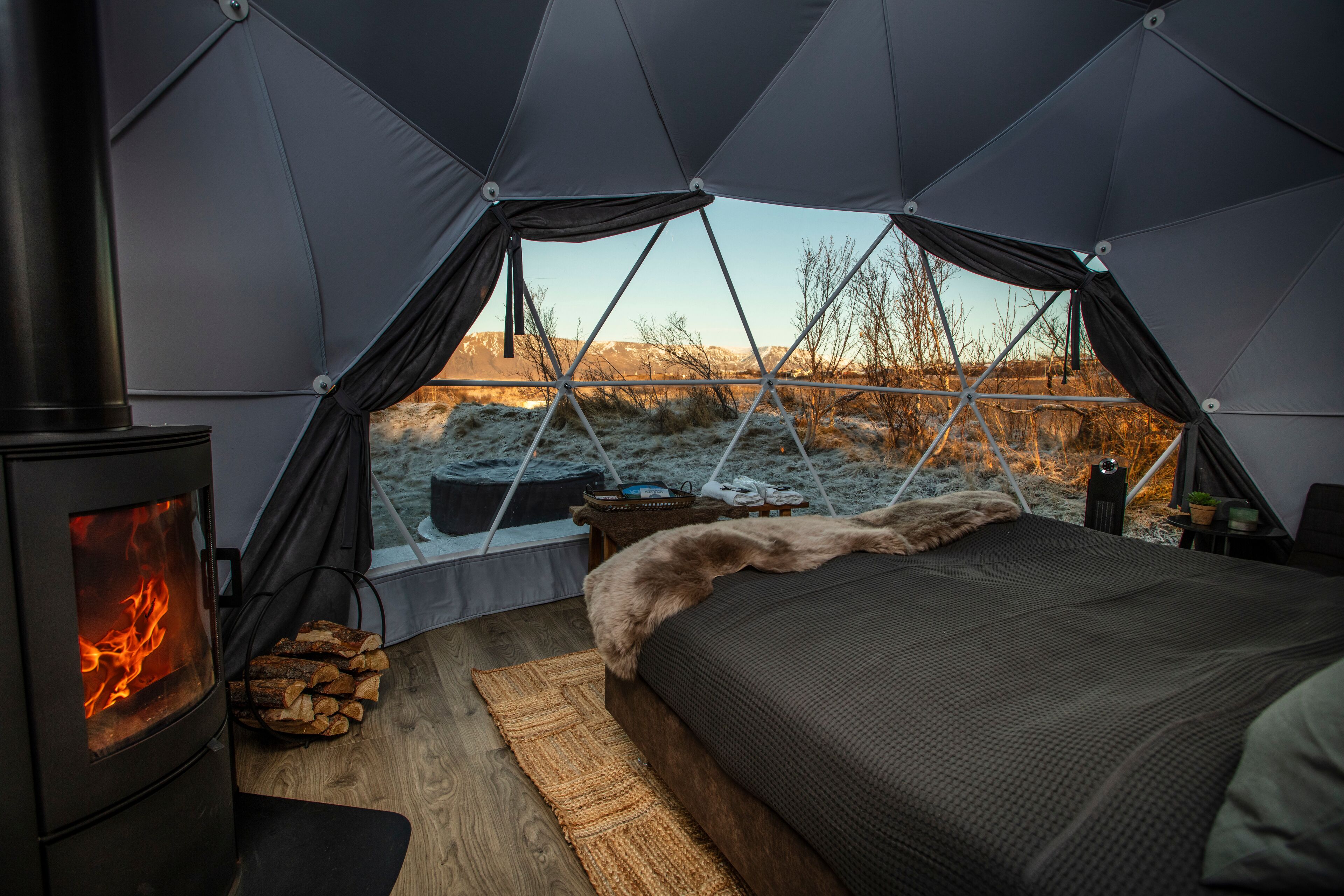 A cozy tented room with a wood-burning stove, a bed, and a view of a snowy landscape.