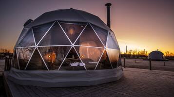 A geodesic dome structure with a transparent roof, a chimney, and a wooden deck in front.