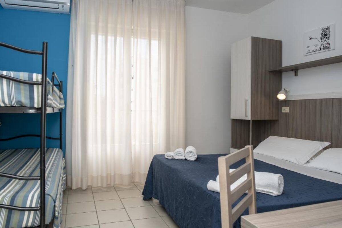 Quarto de hotel com beliche, uma cama de solteiro, escrivaninha, cadeira, janela com cortinas finas e prateleira na parede.