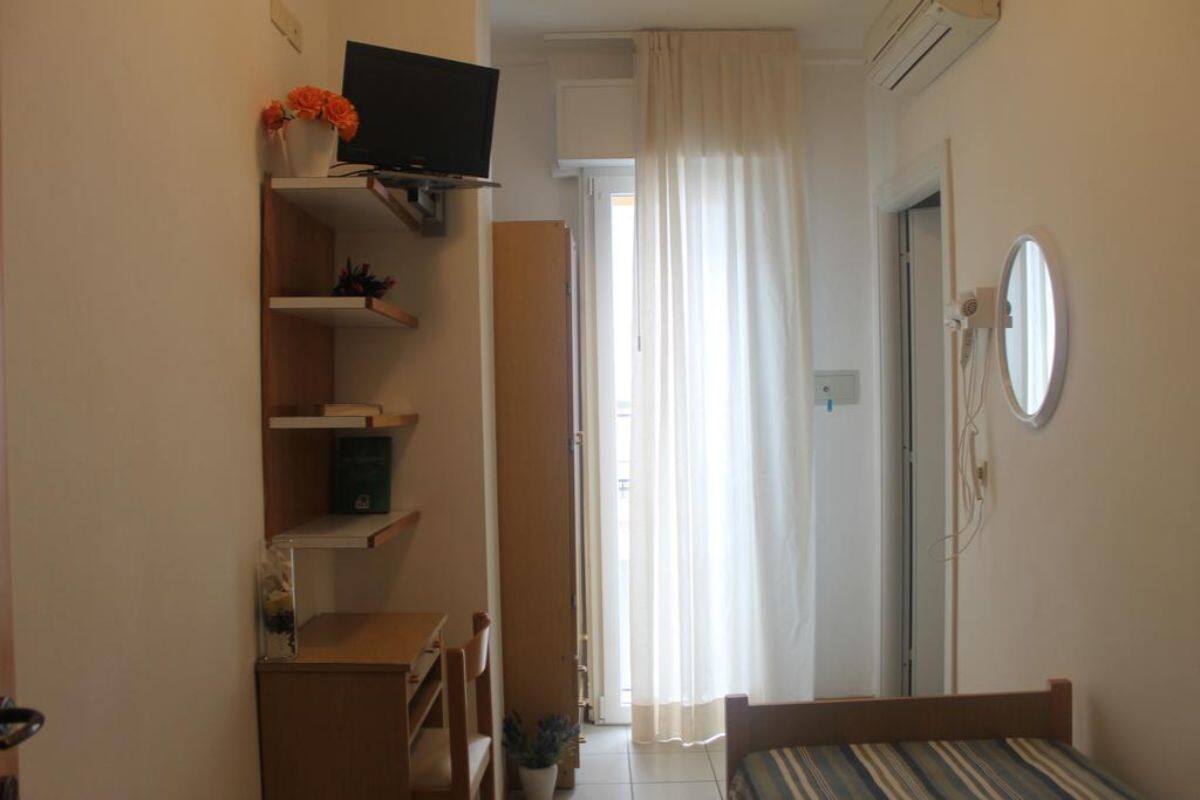 Quarto com cama, televisão numa estante, escrivaninha e janela com cortinas.