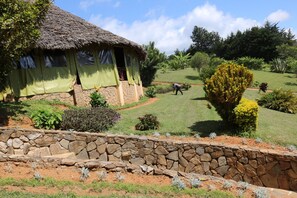 A thatched-roof hut with a stone base, surrounded by a landscaped garden with various plants and a stone retaining wall. - Taita Rocks Hotel (Taita Hills)