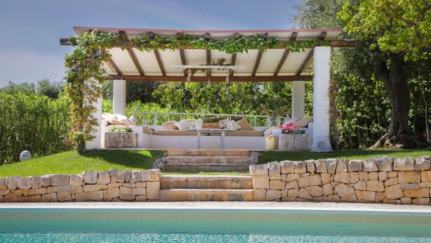 A poolside area with a pergola, outdoor seating, and a stone wall.