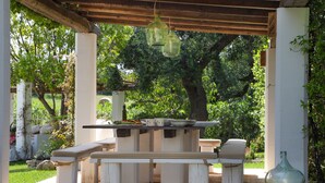 An outdoor patio with a wooden table and benches, surrounded by greenery and trees.