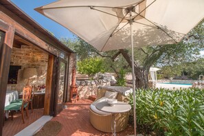 A patio area with a wicker sofa, a table, and a large umbrella. There is a stone wall and a pool in the background. - I Trulli di Annetta (Castellana Grotte)