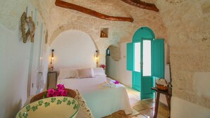 A rustic bedroom with a stone wall, a wooden ceiling beam, a bed with white linens, a small table with a basket, and a teal door. - Il Pinnacolo Verde (Alberobello)