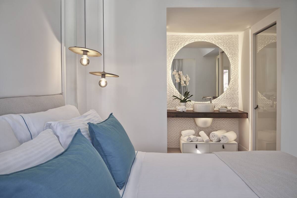 Une chambre à coucher moderne avec un lit, des oreillers, une table de chevet et une commode. Un miroir est accroché au-dessus de la commode et une petite plante en pot est posée sur la table de chevet.