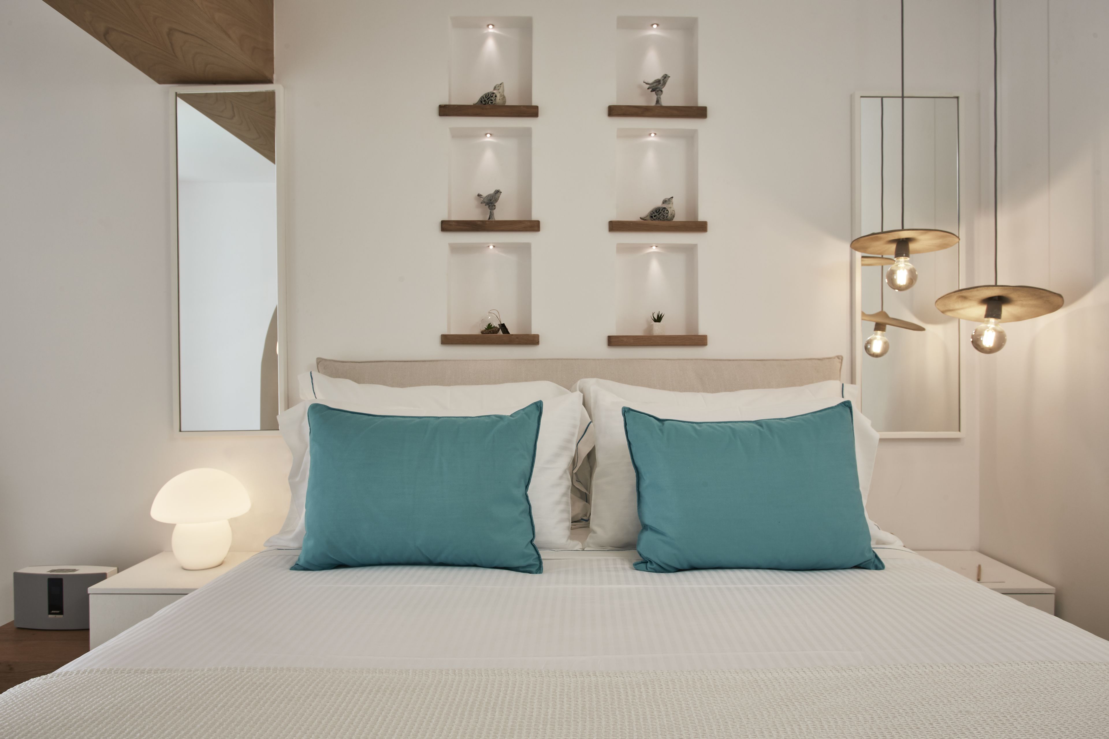 Une chambre moderne avec un grand lit, deux coussins turquoise, une table de chevet avec une lampe et des étagères murales ornées d’objets décoratifs.