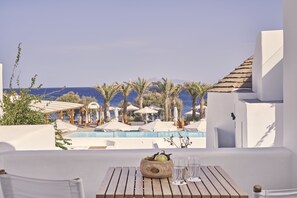 Terraço com mesa de madeira, uma tigela de frutas e copos, com vista para uma piscina e palmeiras, e uma paisagem marinha.