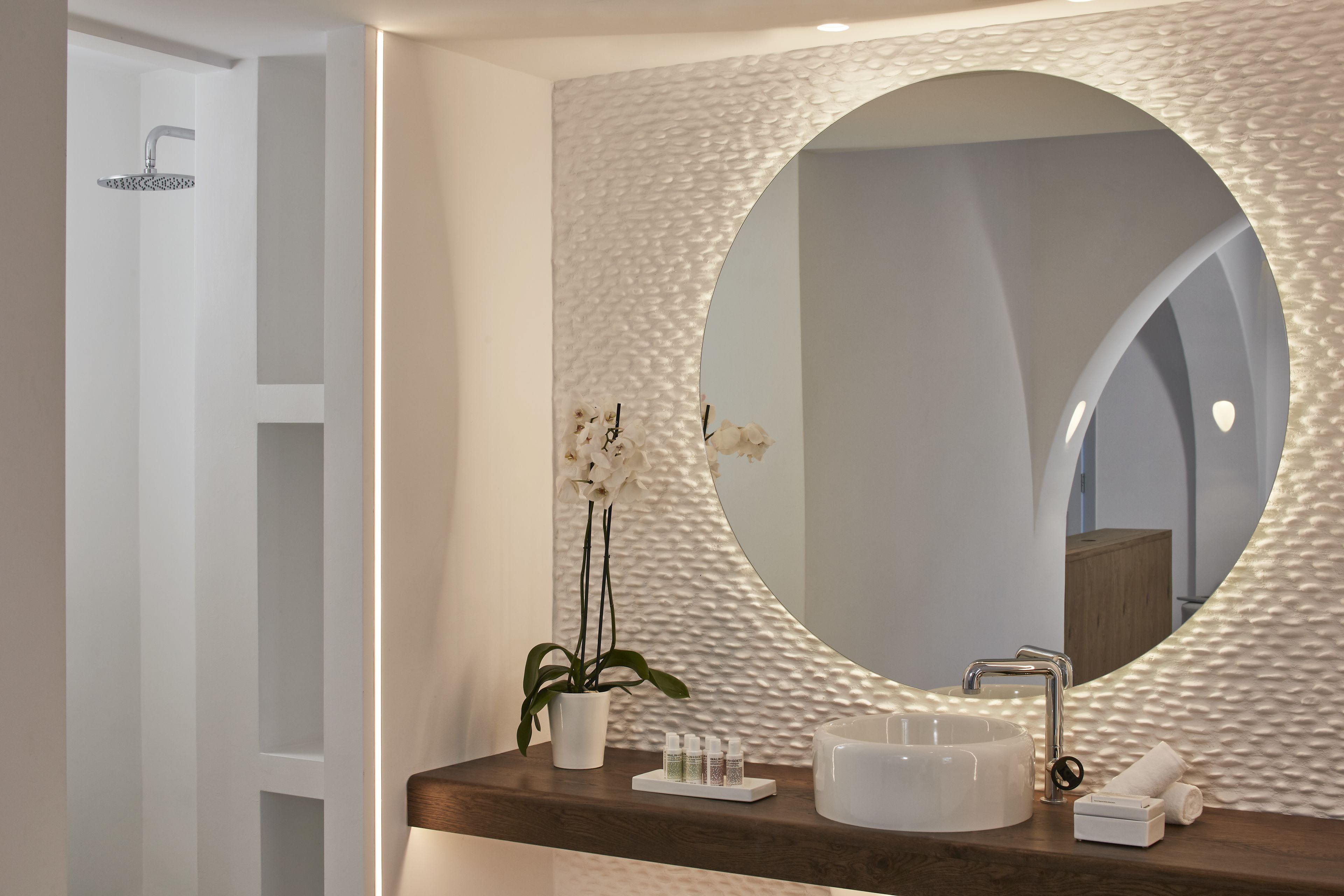 A bathroom with a round mirror, a white sink, a shower, and a potted orchid.