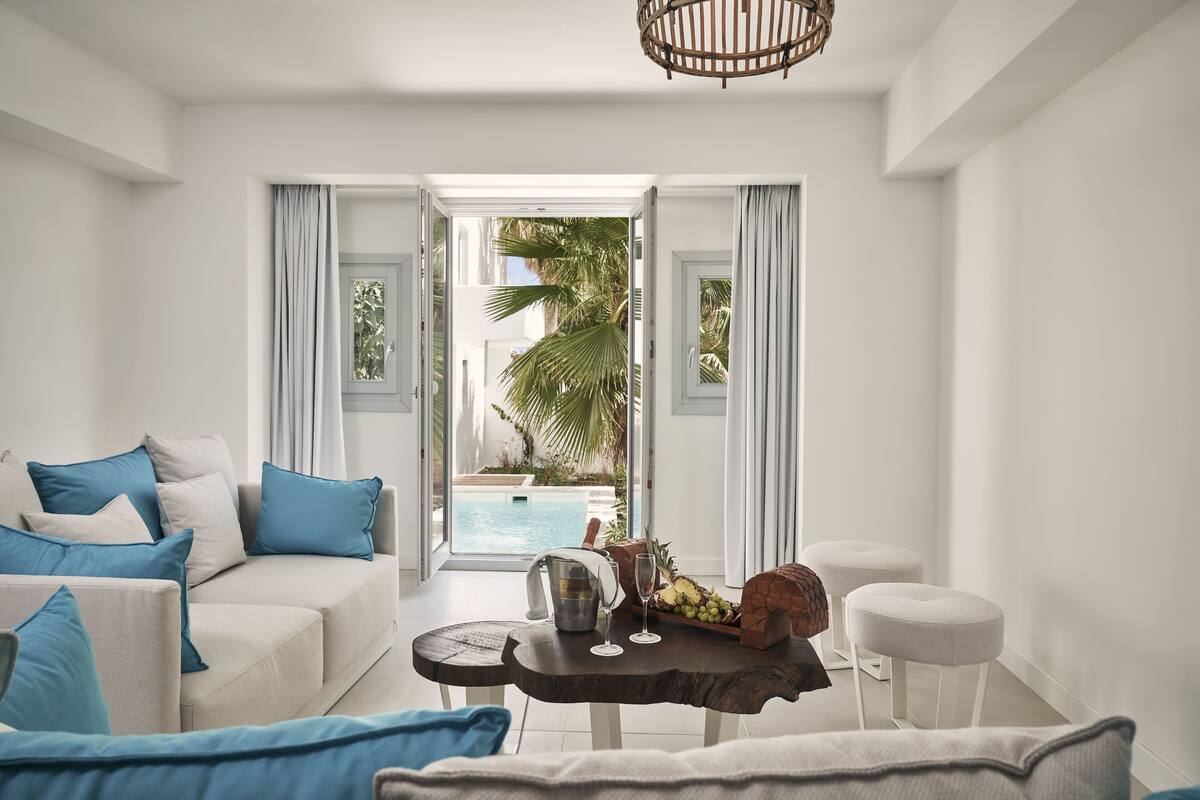 Un salon moderne avec un canapé beige, des coussins bleus, une table basse sur laquelle se trouve un panier de fruits, et une vue sur une piscine et des palmiers.