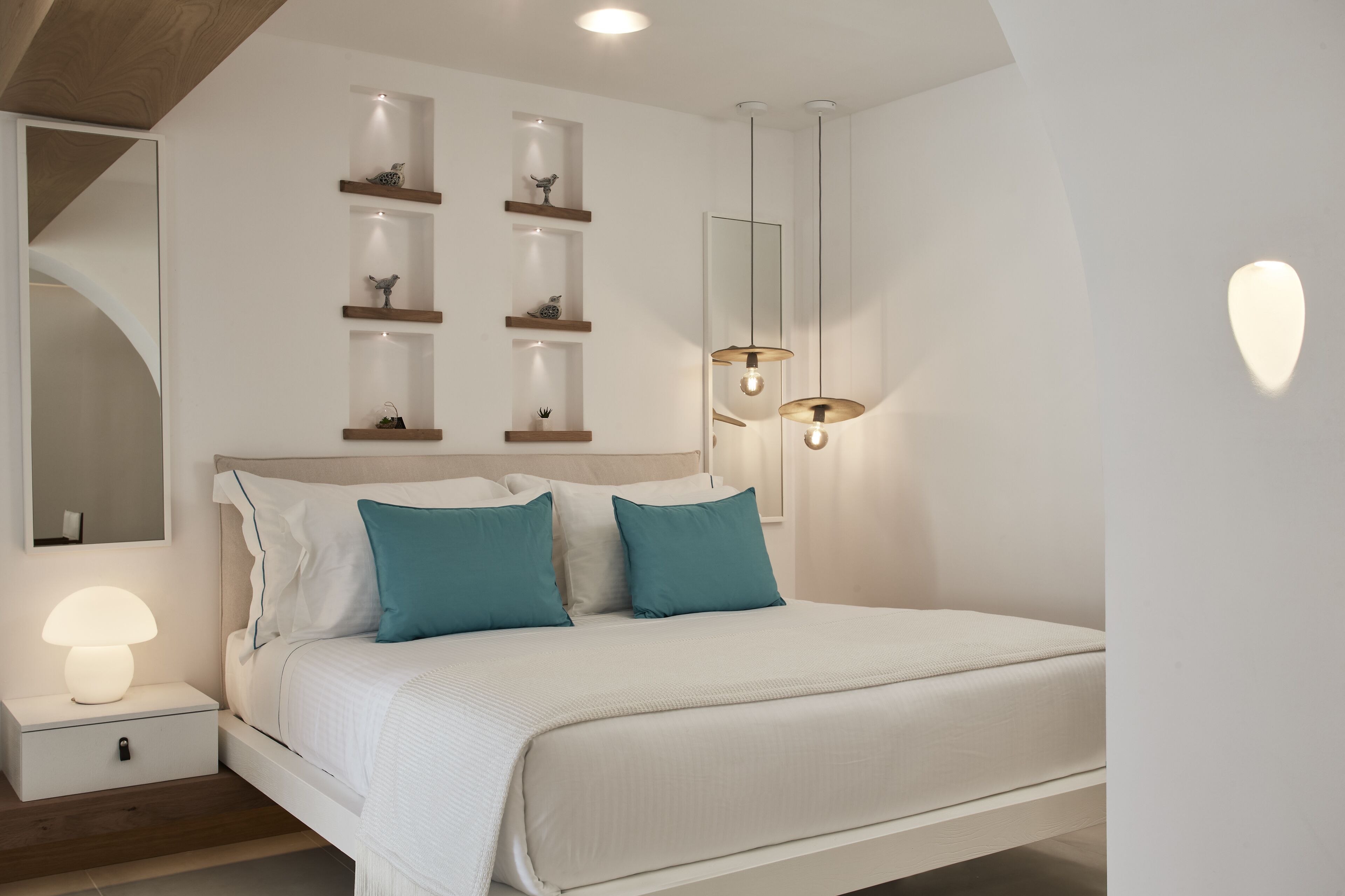 A modern bedroom with a bed, bedside tables, a mirror, shelves with decorative items, and pendant lights.