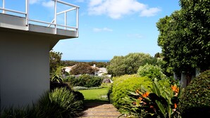 A garden pathway leading to a white building with a balcony overlooking a lush landscape and the sea. - Home with an Ocean view (Hermanus)