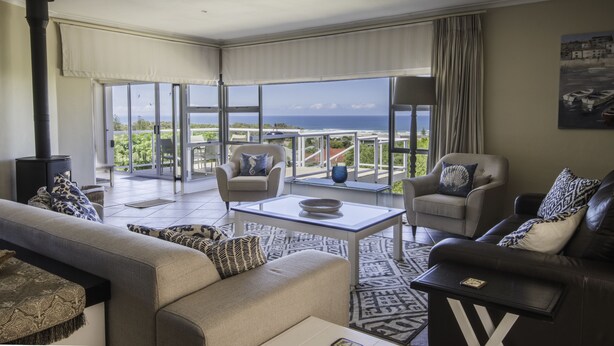 A spacious living room with a large glass door leading to a balcony with a view of the ocean. There are two sofas, a coffee table, and a fireplace. - Home with an Ocean view (Hermanus)