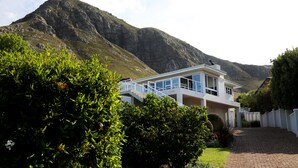 A modern two-story house with a balcony, surrounded by lush greenery and a mountain backdrop. - Home with an Ocean view (Hermanus)