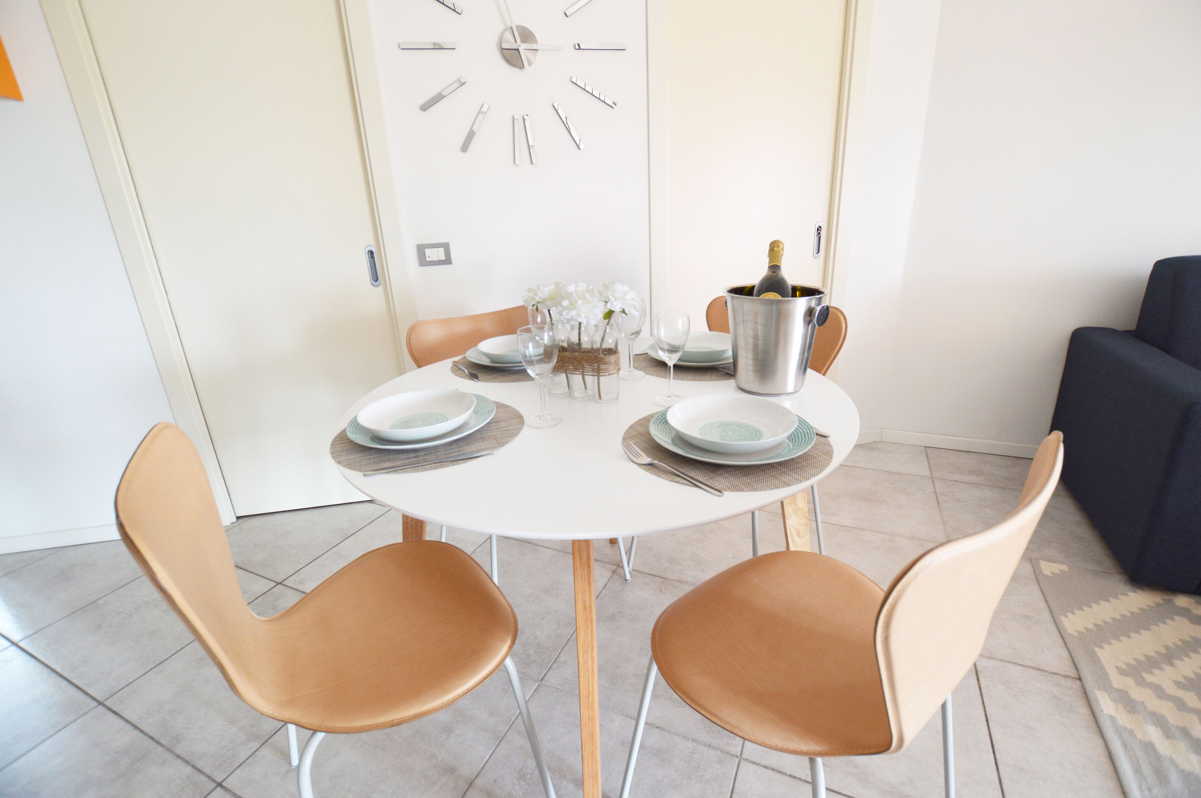 A dining area with a round table, two chairs, a clock on the wall, and a bottle of champagne.