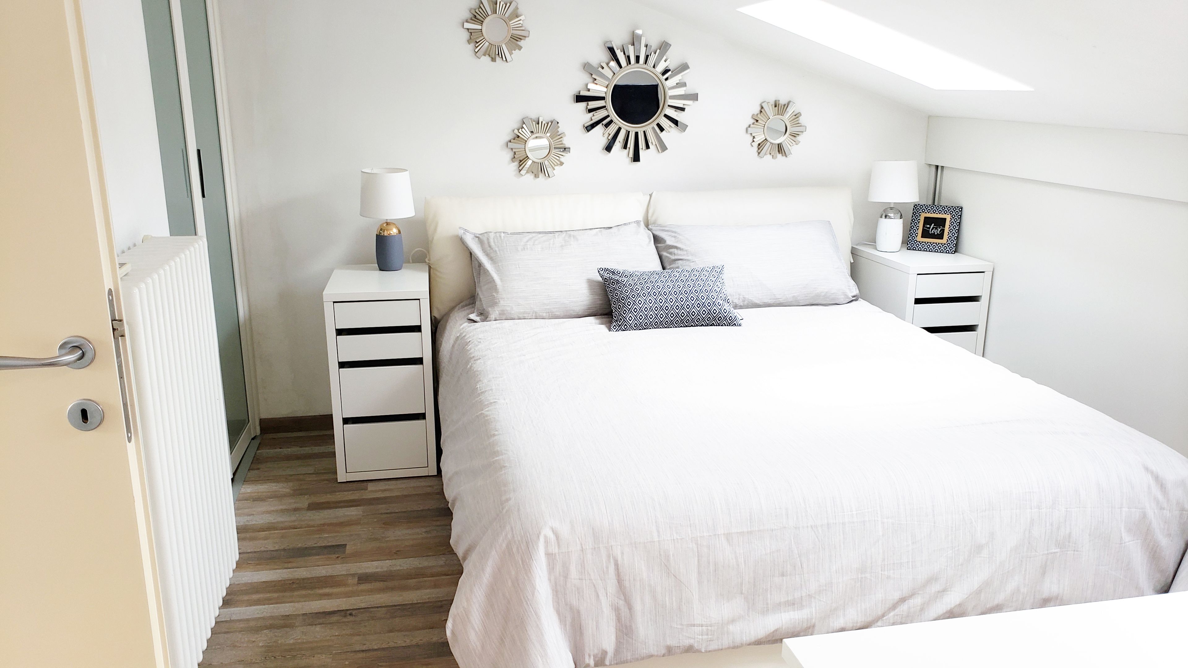 Un dormitorio con una cama grande, dos mesitas de noche, un radiador y espejos decorativos en la pared.