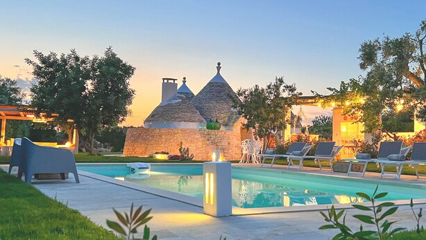 Un área de piscina con sillones de descanso, un edificio de piedra y una pérgola con luces colgantes.