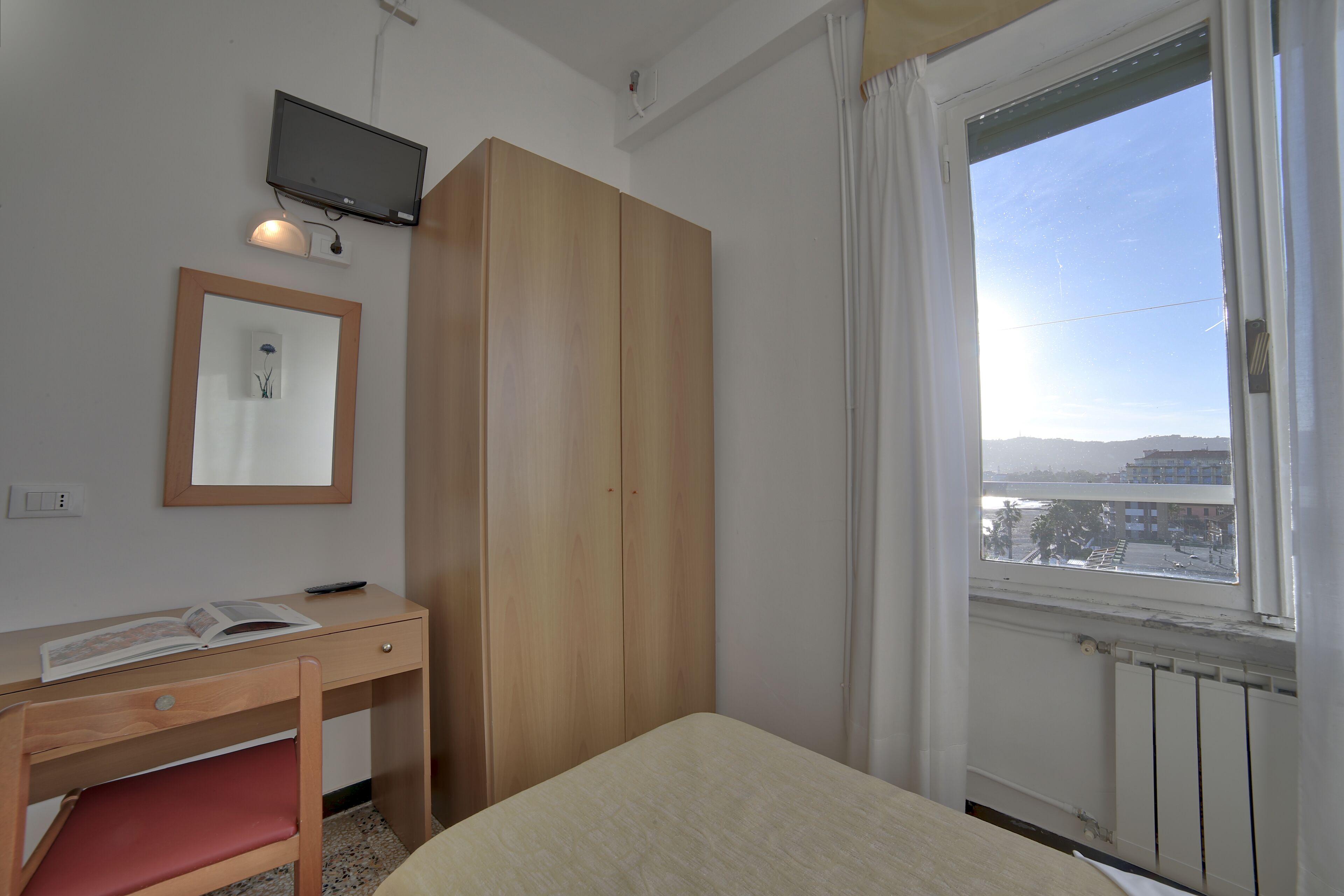 A room with a bed, a desk with a chair, a wardrobe, a television mounted on the wall, and a window with a view of buildings.