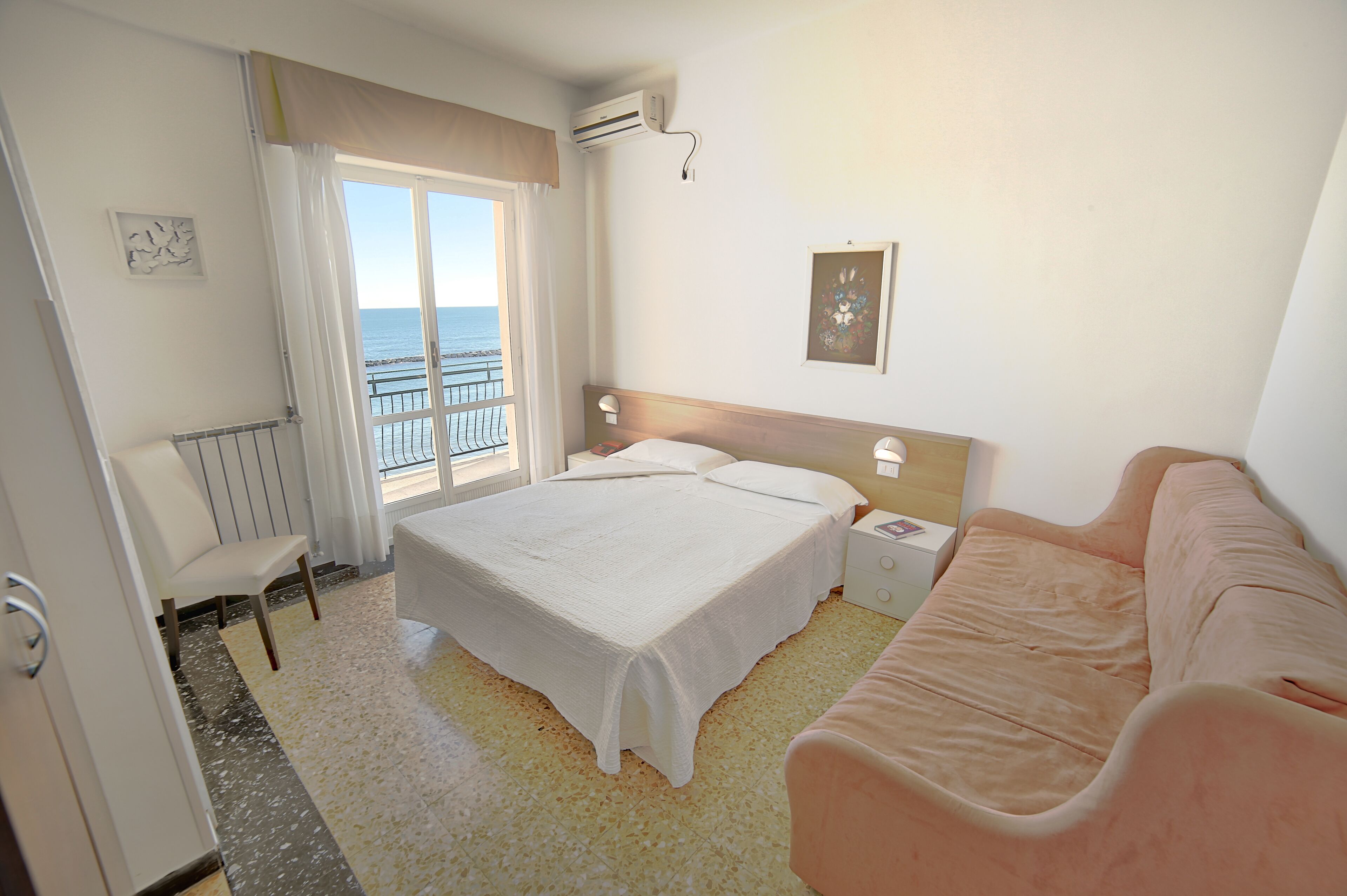 Una camera d'albergo con un letto, una sedia, un comodino e vista mare attraverso una finestra.