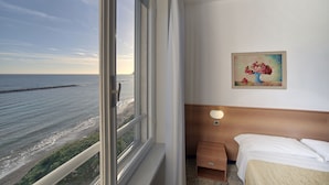 Habitación de hotel con un ventanal grande que da a una playa, un cuadro en la pared y una cama con cabecero de madera.
