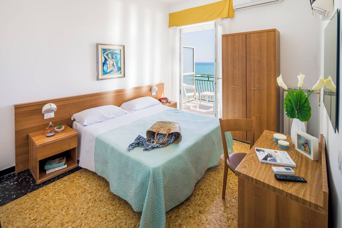 Een hotelkamer met een bed, een bureau, een stoel, een nachtkastje, een kledingkast, een raam met uitzicht op zee en een plant.