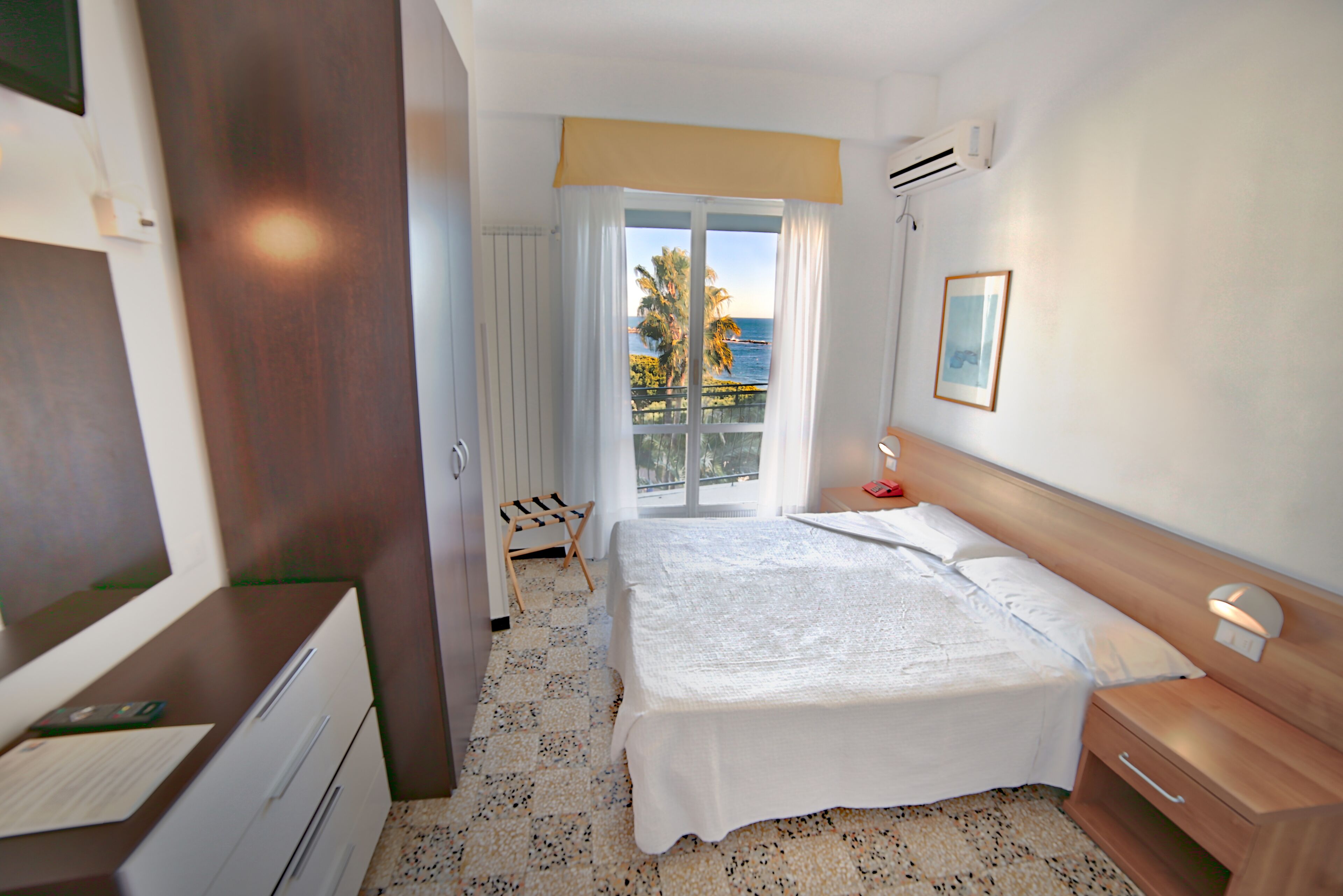 Habitación de hotel con cama, escritorio, silla, ventana con vistas a palmeras y al mar, y unidad de aire acondicionado.